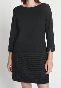 Svart klänning med långa ärmar, med en figurnära överdel och en strukturerad, vadderad nederkant. Ärmarna har en liten slits och manschettdetalj.