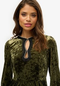 Robe en velours vert avec des manches longues, dotée d'un décolleté en forme de clé noir orné d'ornements en argent. Texture douce et design élégant.