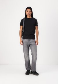 T-shirt noir à manches courtes, jeans gris à coupe droite et baskets noires avec des accents blancs. L'arrière-plan est blanc uni.