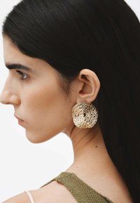 Aretes de oro tejidos en forma redonda con un diseño texturizado y parecido a una malla. El modelo los muestra puestos, resaltando su tamaño y su brillo metálico.