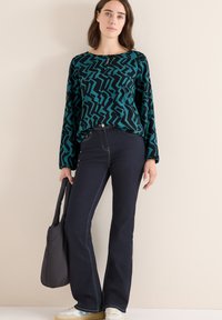 Mørkeblå og sort mønstrede langærmede top med rund halsudskæring; parret med mørke jeans og afslappede sneakers; holder en mørk tote bag.