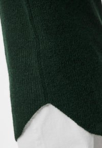 Maglione a maglia verde scuro con una finitura testurizzata. Presenta un orlo frastagliato, che mette in mostra un indumento bianco a contrasto sottostante.