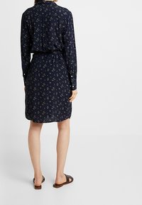 Granatowa sukienka koszulowa z długim rękawem i dopasowaną talią, z małym kwiatowym wzorem. Nosić z brązowymi sandałami, gładka tekstura materiału.