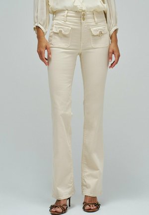 Mujer con pantalones rectos color crema con dobles bolsillos delanteros con solapa y cierre de botones, combinados con tacones negros de tiras.