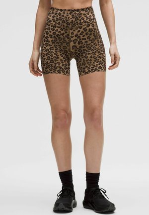 Žena v atletických kraťasoch s vysokým pásom so vzorom leopardieho tigrieho vzoru, v kombinácii s čiernymi ponožkami a čiernymi bežeckými topánkami, stojaca na jednoliatom pozadí.