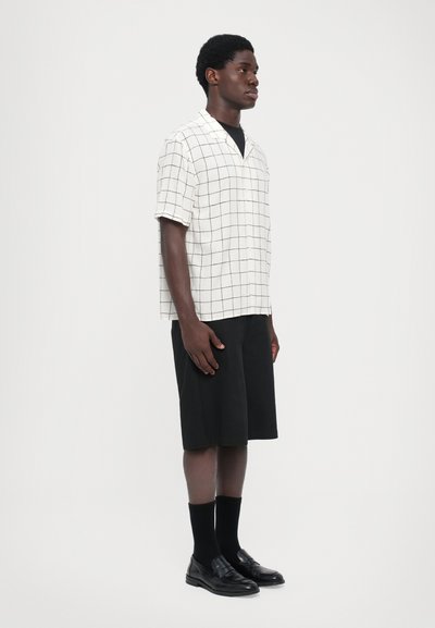 Korte mouwen overhemd met kraag in crèmekleurige stof met een zwart ruitpatroon, gecombineerd met zwarte shorts en zwarte instappers met sokken.