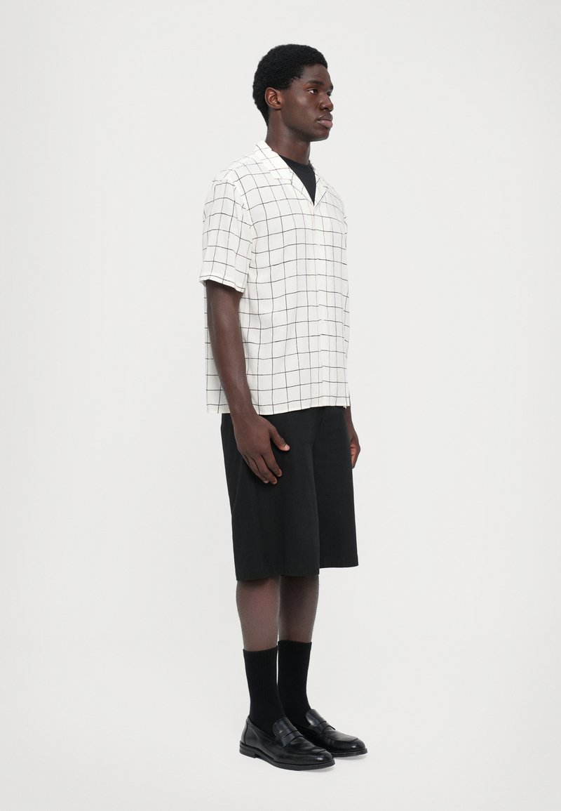 Kortärmad skjorta med krage i krämfärgad tyg med svart rutmönster, kombinerad med svarta shorts och svarta slippers med strumpor.