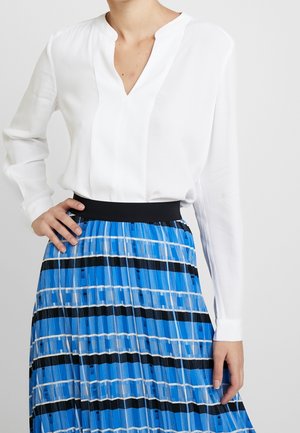 Nainen, joka pitää valkoista pitkähihnaista paitapuseroa, joka on sullottu korkeavyötäröiseen sinivalko-mustaraidalliseen laskostettuun hameeseen.