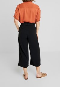 Chemise à manches courtes de couleur rouille avec une coupe ample, associée à un pantalon large noir à taille haute et poches, portée avec des sandales beiges.