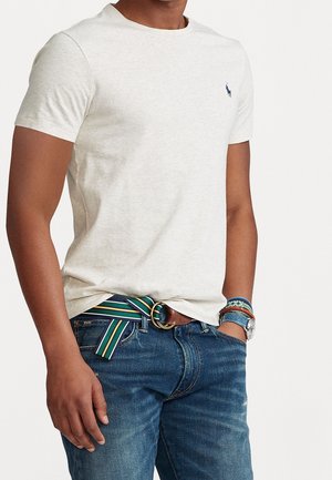 Personne portant un t-shirt beige clair ajusté avec un petit logo bleu marine, un jean bleu, une ceinture rayée verte et jaune, ainsi que des bracelets et une montre au poignet.