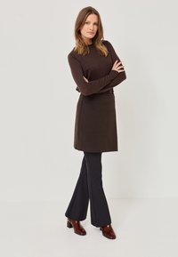 Top lung cu maneci lungi de culoare maro, asortat cu o fustă maro închis și pantaloni evazați negri. Modelul poartă botine cu toc lucios de culoare maro. Design simplu.