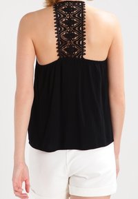 Svart ärmlös topp med en strukturerad, lös passform. Har en spetsbak med intrikata mönster. Bärs med vita shorts.