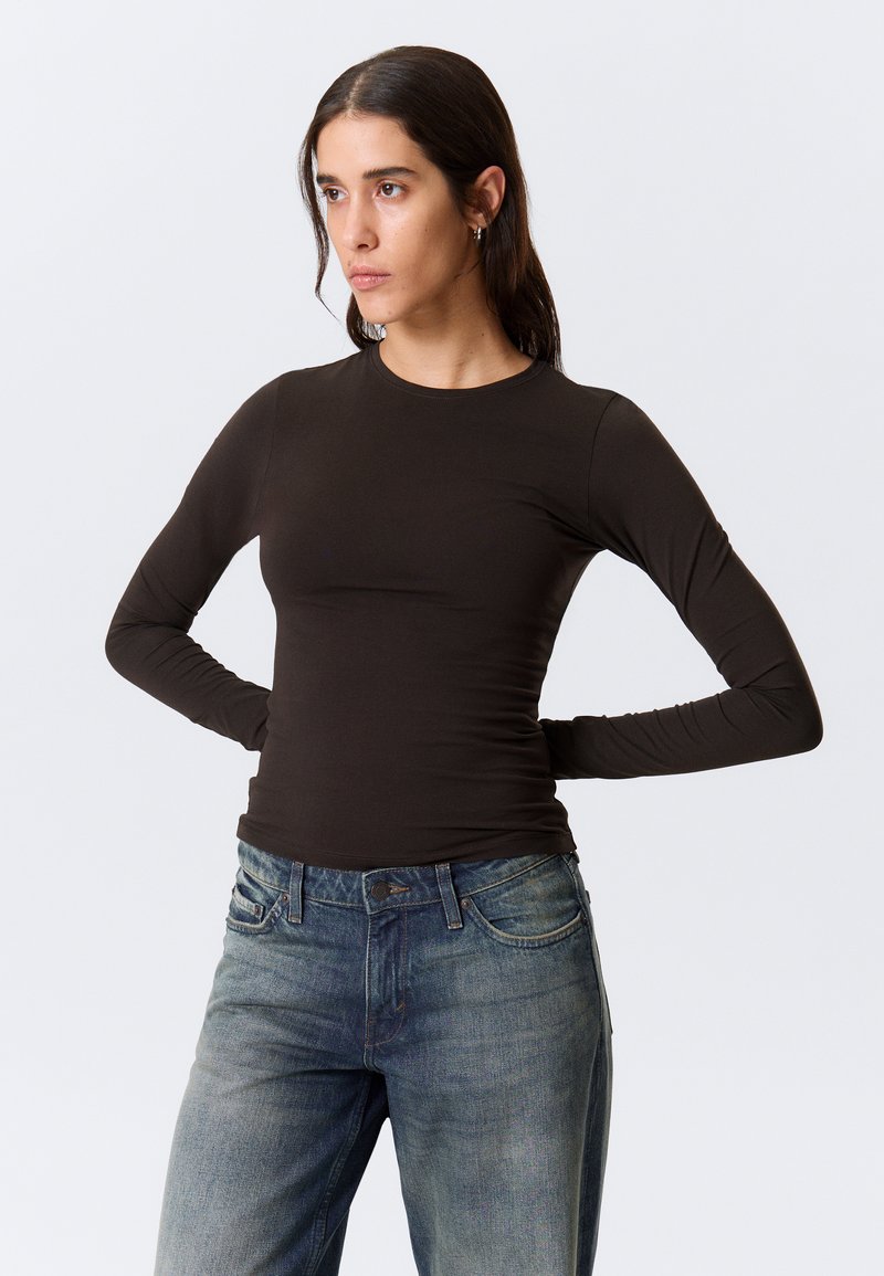 Long-sleeve brown top made of stretchy fabric, featuring a round neckline. Paired with medium-wash denim jeans.