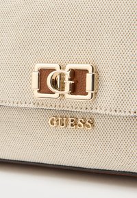 Beige texturált külsővel rendelkező vászon kézitáska. Arany csattal és barna bőr részlettel, amelyen arany "GUESS" logó található.