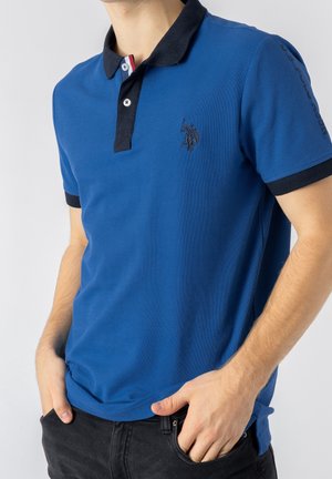 Uomo che indossa una polo blu con colletto scuro, logo ricamato sul petto e testo di marca sulla manica, mani nelle tasche dei jeans neri.