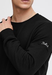 Zwarte shirt met lange mouwen van katoen, met een ronde hals en geribde manchetten. Heeft een textuurpatch op de linkermouw met het woord "Solid."