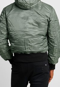Persona che indossa un giubbotto imbottito verde con polsini e orlo nero a costine, abbinato a pantaloni neri, visto da dietro.