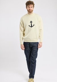 Pull en tricot crème avec un motif d'ancre noir sur le devant, associé à un jean en denim foncé. Le mannequin porte des chaussures beiges.