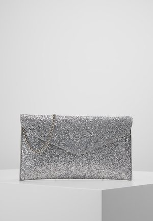 Sølvglitter kuvertclutch med en tynd sølvkæderem på en hvid piedestal mod en ensfarvet grå baggrund.