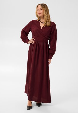 Femme portant une robe longue bordeaux à manches longues, avec un col en V et une taille froncée, debout avec une main sur la hanche.