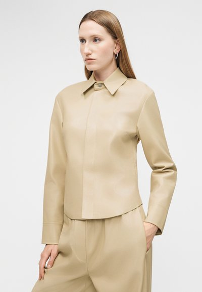 Žena s rovnými blond vlasmi nosí béžovú koženú bundu na gombíky a zodpovedajúce nohavice, stojaca s jednou rukou v vrecku na jednoduchom pozadí.