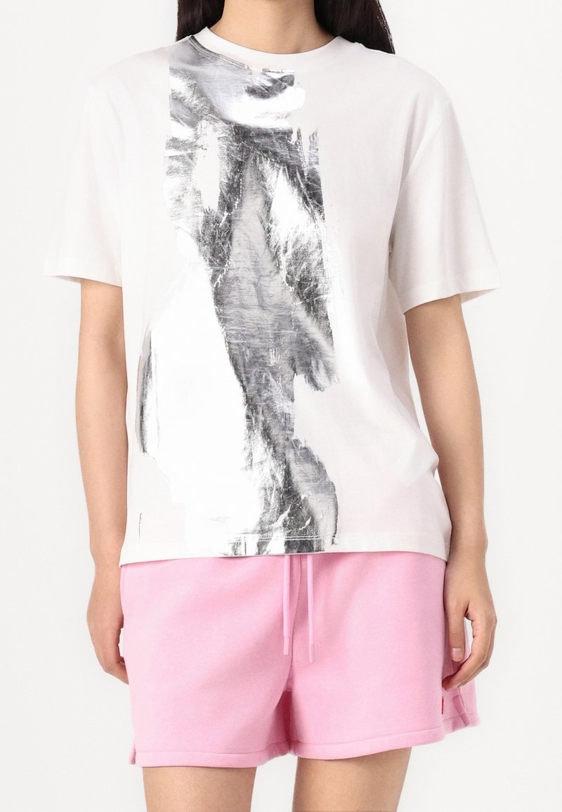 Femme portant un t-shirt blanc avec un motif abstrait argenté et un short rose avec cordon de serrage sur un fond uni.