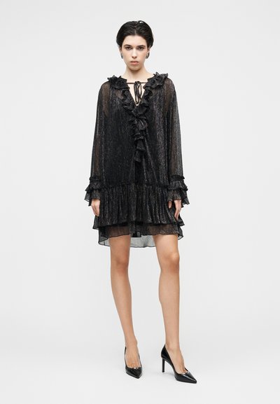 Μαύρο, λαμπερό φόρεμα με βολάν στον λαιμό και τα μανίκια, με στρώσεις στο τελείωμα, που δένει μπροστά. Φορεμένο με μαύρες γόβες.