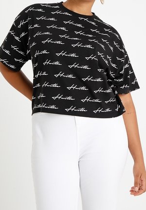 Persona vistiendo una camiseta negra recortada con la palabra "Hustle" repetida en blanco y pantalones blancos de talle alto, de pie con una mano en la cadera.