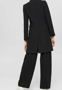 Person, der einen langen schwarzen Mantel mit einem Rückenschlitz, weiten schwarzen Hosen und weißen Schuhen trägt, steht vor einem hellgrauen Hintergrund.