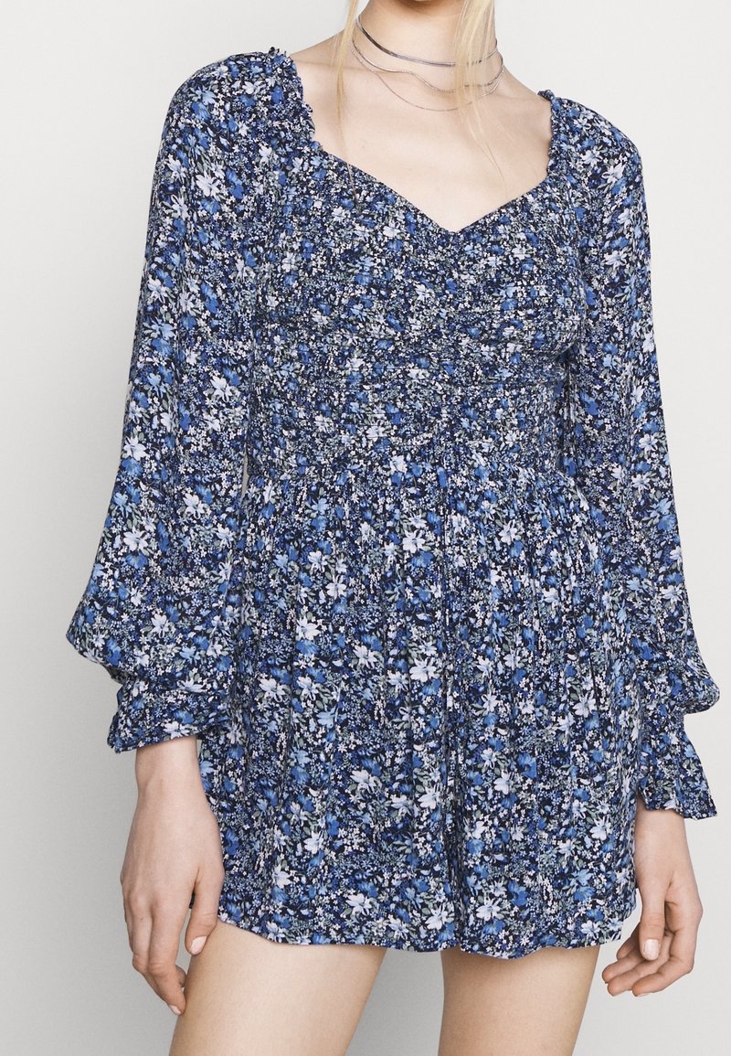 Femme portant une robe longue à manches longues bleue à fleurs, avec un corsage froncé et un décolleté en cœur, debout devant un fond uni.