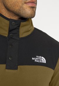 Jersey de forro polar en negro y verde oliva, con cuello alto, parte delantera abotonada y logo bordado. Textura suave, diseño sencillo.