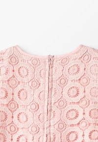 Ljusrosa spets tyg med cirkulära blommönster och en dragkedja med pärlhandtag vid halslinningen på ett plagg.