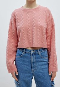 Rosa, kabelgestrickter, kurz geschnittener Pullover mit langen Ärmeln und strukturiertem Design, kombiniert mit hochgeschnittenen blauen Jeans.
