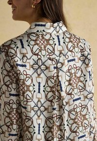 Silky shirt featuring a complex geometric pattern with equestrian motifs in brown, green, blue, and beige on a light background.