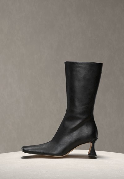 Μαύρη δερμάτινη μπότα μέχρι τη μέση της γάμπας με μυτερή μύτη και καμπύλο τακούνι στιλέτο, εμφανίζεται σε ουδέτερο φόντο πάνω σε φωτεινή επιφάνεια.