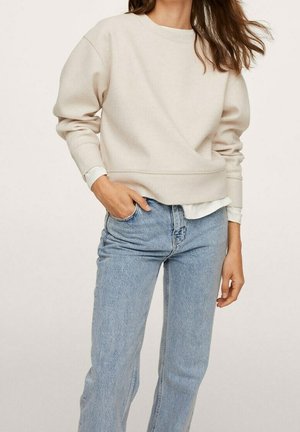 Femme portant un sweat-shirt beige à col rond par-dessus une chemise blanche, associé à un jean taille haute bleu clair, debout avec une main dans la poche.