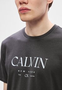 Hallides hall t-särk, millel on suur valge "CALVIN" trükk, väiksemad tekstid "NEW YORK," "1968," ja "CK." Ümar kaelus ja lühikesed varrukad.