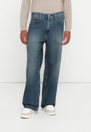 Homme portant un jean en denim bleu ample, une chemise beige à manches longues et des baskets blanches, debout contre un fond blanc uni.