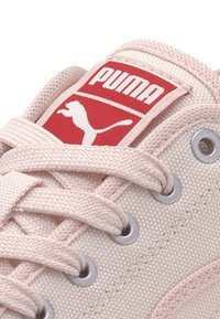 Gros plan d'une basket Puma rose clair avec des lacets en tissu passés dans des œillets métalliques et un logo Puma rouge sur la languette.