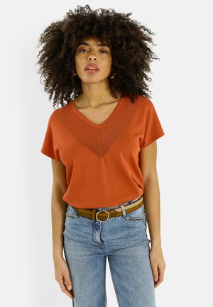 Femme aux cheveux bouclés portant un haut orange à col en V et manches courtes, un jean bleu clair et une ceinture marron avec une boucle ronde, sur fond blanc.