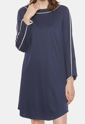 Donna che indossa un vestito blu navy a maniche lunghe con bordatura bianca su maniche e scollo, mano appoggiata vicino alla clavicola.
