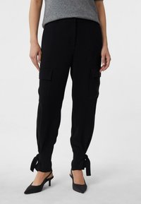 Czarne spodnie cargo mają boczne kieszenie, zwężane nogawki i kokardki na mankietach. Materiał wydaje się miękki i o gładkiej fakturze.