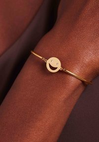 Gouden armband met een glimlachend gezicht charme en twee heldere stenen accenten, geplaatst tegen een textuur van huid.