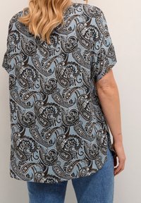 Blusa de corte suelto en azul claro con un patrón de paisley negro. Mangas cortas, dobladillo redondeado y tela texturizada.