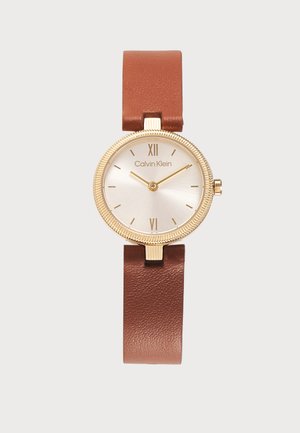 Reloj redondo de oro con esfera blanca, correa de cuero marrón, números romanos en 12 y 6, y logotipo de Calvin Klein en la esfera.