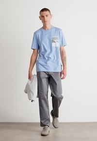 Lyseblå kortermet T-skjorte med lomme som har et grått mønster, kombinert med grå bukser og beige sneakers.