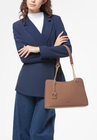 Borsa a tracolla marrone con texture e dettagli in catena dorata, con forma rettangolare e cartellino pendente. Indossata con un blazer blu navy e pantaloni in denim.