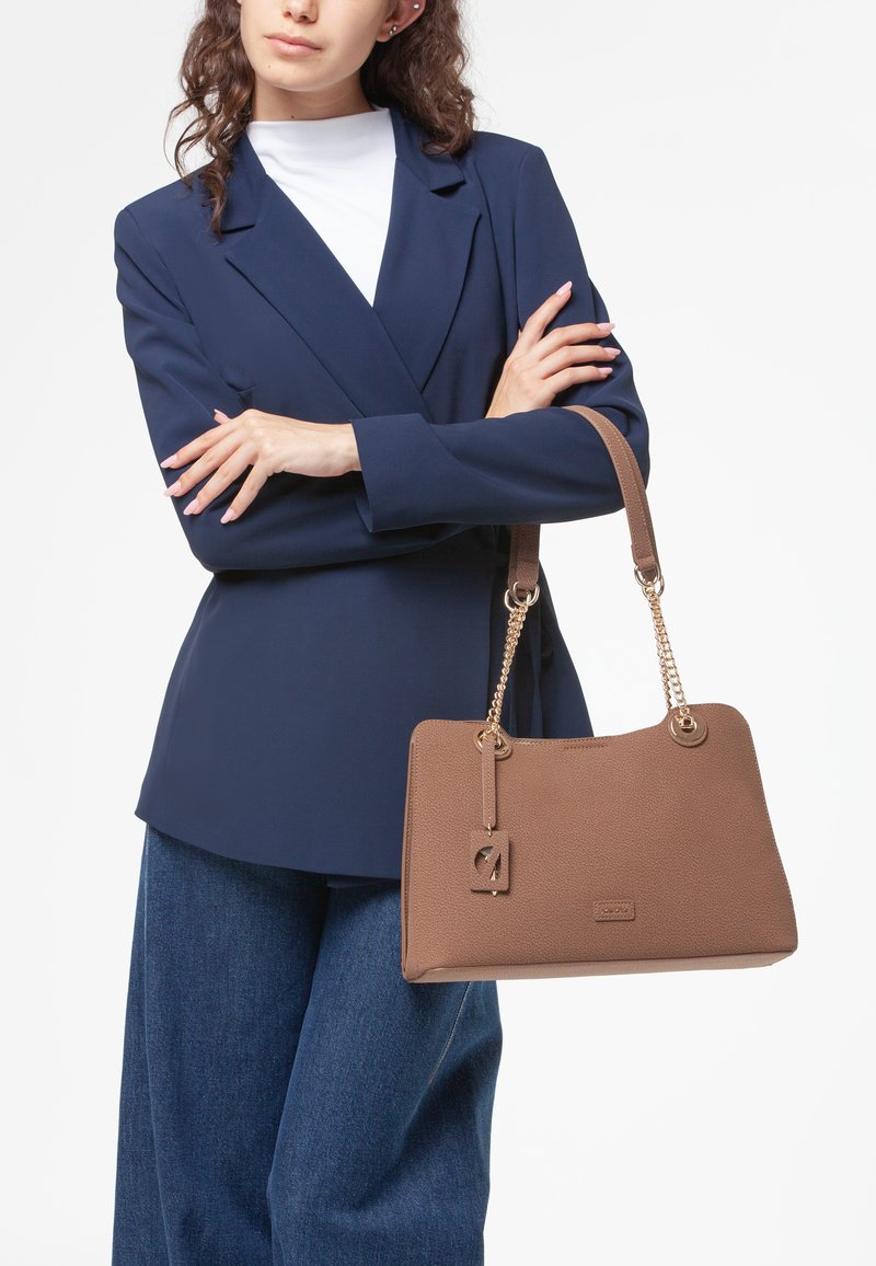 Borsa a tracolla marrone con texture e dettagli in catena dorata, con forma rettangolare e cartellino pendente. Indossata con un blazer blu navy e pantaloni in denim.