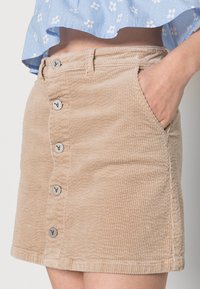 Saia de veludo cotelê em tom de bege, com riscas verticais, bolsos frontais e fecho de cinco botões com acabamentos em metal. Design casual, até ao joelho.
