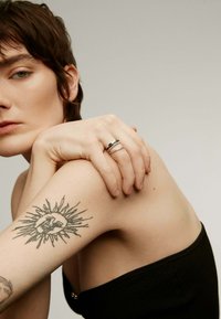 Anillos de plata apilados con acentos negros en una mano reposando sobre un hombro descubierto con un tatuaje de sol, todo contra un fondo tenue.